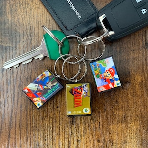 May include: A set of three keychains featuring retro video game cartridge designs. The keychains are made of clear plastic and feature the game titles "Super Mario World", "Yoshi's Island", and "Zelda: A Link to the Past".