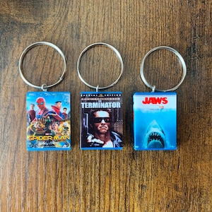 以下が含まれることがあります： 映画をテーマにしたデザインのキーホルダーが3つ。キーホルダーはシルバーで、青い背景に映画のポスターデザインが施されています。キーホルダーは、「スパイダーマン」、「ターミネーター」、「ジョーズ」の映画をフィーチャーしています。