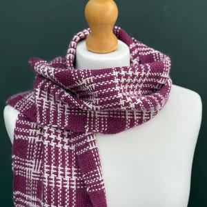 May include: A plum-coloured woven scarf with a white plaid design is displayed on a mannequin. The scarf is made of a soft, textured material. The scarf is wrapped around the mannequin's neck and hangs down the front.