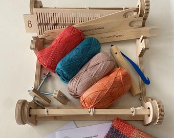May include: A wooden rigid heddle loom with four balls of yarn in red, teal, peach, and orange. The loom is set up with a weaving pattern and a finished woven scarf in a red, teal, peach, and orange plaid pattern.