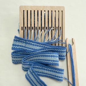 May include: A wooden weaving loom with blue and white woven fabric. The fabric features a blue and white striped pattern. A wooden shuttle with blue thread is also present. The loom is rectangular with vertical slots.