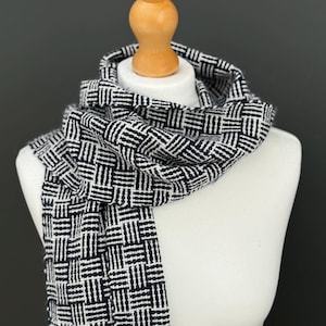 Puede incluir: Una bufanda tejida en blanco y negro con un patrón geométrico está drapeada sobre un maniquí blanco. El diseño de la bufanda presenta una cuadrícula repetida de líneas oscuras y claras, creando una apariencia texturizada. La bufanda se muestra sobre un fondo gris oscuro.