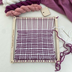 May include: A wooden loom with a woven tapestry in shades of white and purple. The loom has a label that reads "TabbyandTweed". Three skeins of yarn in white, pink, and purple are in the background.