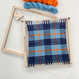 May include: A wooden weaving loom with a blue, orange, and light blue plaid pattern woven on it. Three balls of yarn in the same colors are included.