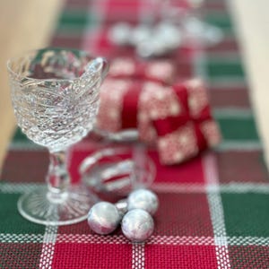 May include: A clear crystal wine glass with a detailed pattern sits on a red and green plaid table runner. Silver ornaments and wrapped gifts with red ribbons are also on the table, creating a festive holiday scene.