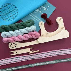 May include: A craft kit with a wooden weaving loom, yarn in shades of green, blue, and pink, and woven ribbons. A blue bag with the text "TabbyandTweed" is in the upper left corner. A card with the text "Thank You" is in the upper right corner.