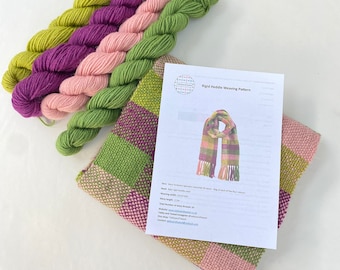 Op de afbeelding: Een weefset voor een sjaal met vier garenklossen in groen, roze en paars. De set bevat een patroon voor een starre weefgetouw, met instructies voor het weven van een sjaal. Het patroon bevat de tekst "Rigid Heddle Weaving Pattern".