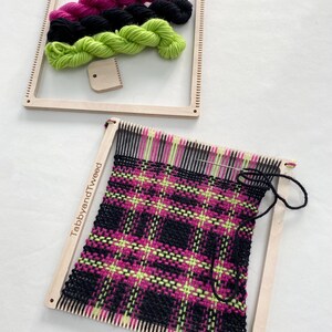 May include: A wooden loom with a pink, black, and green plaid pattern woven on it. The loom is labeled "TabbyandTweed". There are three balls of yarn in the background: pink, black, and green.