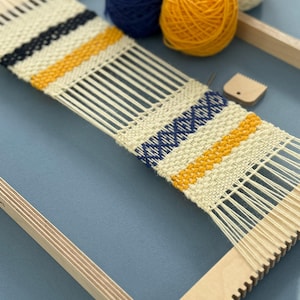 May include: A wooden frame loom with a partially woven textile featuring cream, navy blue, and mustard yellow yarn. The woven piece displays horizontal stripes and a cross-stitch pattern. Three balls of yarn and a small wooden weaving comb are included.