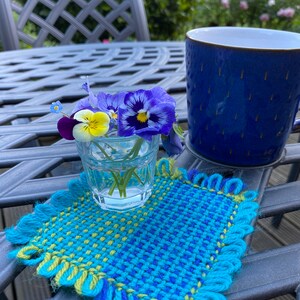 Fabric square woven on a pin less pin loom being used as a coaster for a small vase of flowers.