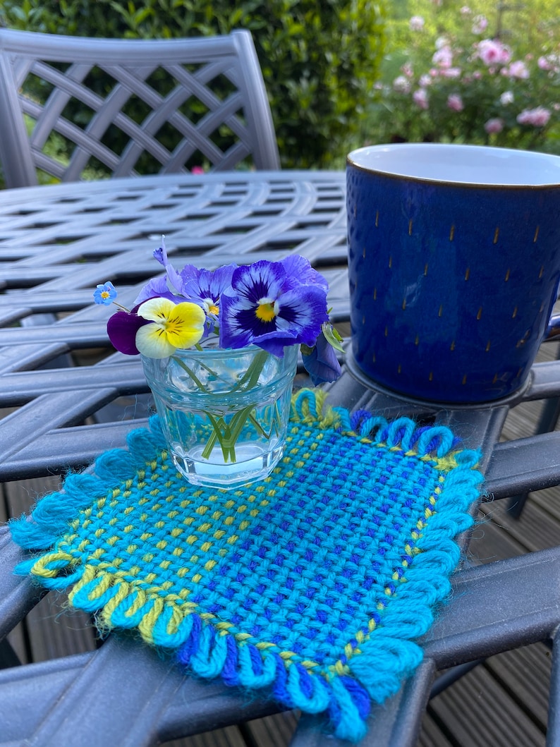 Fabric square woven on a pin less pin loom being used as a coaster for a small vase of flowers.