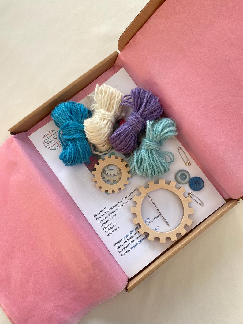May include: A craft kit for making two flower brooches. The kit includes yarn in four colors, two wooden flower frames, a tapestry needle, two buttons, two safety pins, and instructions. The kit is packaged in a pink and brown box.