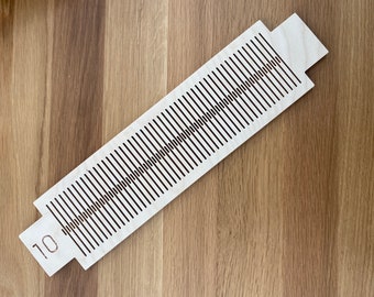 Op de afbeelding: Een lichtgekleurde houten weefgereedschap met een reeks parallelle zwarte lijnen en het nummer 10 gegraveerd aan één uiteinde. Het gereedschap is rechthoekig met verlengde lipjes. De achtergrond is een houten oppervlak.