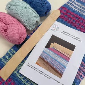 May include: A craft kit featuring a woven textile in blue, pink, and green, with three yarn skeins in pink, blue, and light blue. A wooden stick and a booklet titled "Rigid Heddle Weaving Pattern" are also present. The booklet text reads "Learning to weave with pick up sticks."