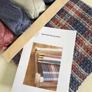 May include: A collection of weaving supplies including yarn, a wooden weaving tool, and a woven fabric sample. The yarn is in shades of blue, gray, and pink. A printed pattern for rigid heddle weaving is also included.