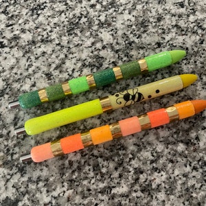 May include: Four glitter pens with gold accents. The pens are green, yellow, orange, and pink. The yellow pen has a bee design.