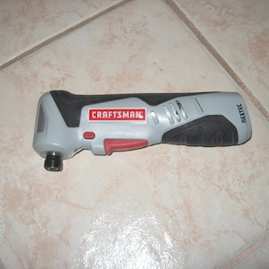 May include: A Craftsman Nextec multi-tool, primarily gray and black, with a red button and the Craftsman logo. The tool is designed for various cutting and sanding tasks. The tool is lying on a light-colored tiled surface.
