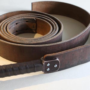 May include: A rolled-up, dark brown leather belt with a matching handle. The belt is approximately 3.8 cm wide. The handle is wrapped in dark brown leather and secured with rivets. The belt is designed for use.