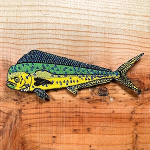 May include: Embroidered patch featuring a colorful mahi-mahi fish. The fish has a yellow body with blue and green accents, and a detailed design. The patch is placed on a wooden surface.