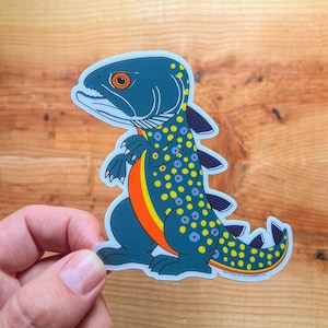May include: A sticker featuring a stylized fish-dinosaur hybrid. The creature is primarily teal with yellow, orange, and blue accents. It has a dinosaur-like posture and fins, with a fish-like head and spotted pattern. The sticker is held by a hand.