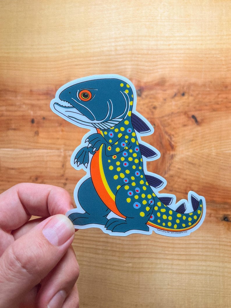 May include: A sticker featuring a stylized fish-dinosaur hybrid. The creature is primarily teal with yellow, orange, and blue accents. It has a dinosaur-like posture and fins, with a fish-like head and spotted pattern. The sticker is held by a hand.