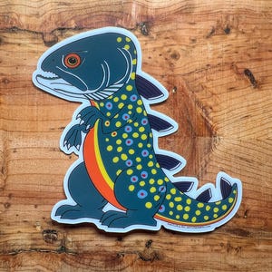 May include: A sticker featuring a cartoon fish-dinosaur hybrid. The creature has a dark teal body with yellow and blue spots, an orange stripe, and a fish-like head. The sticker is on a wooden surface.