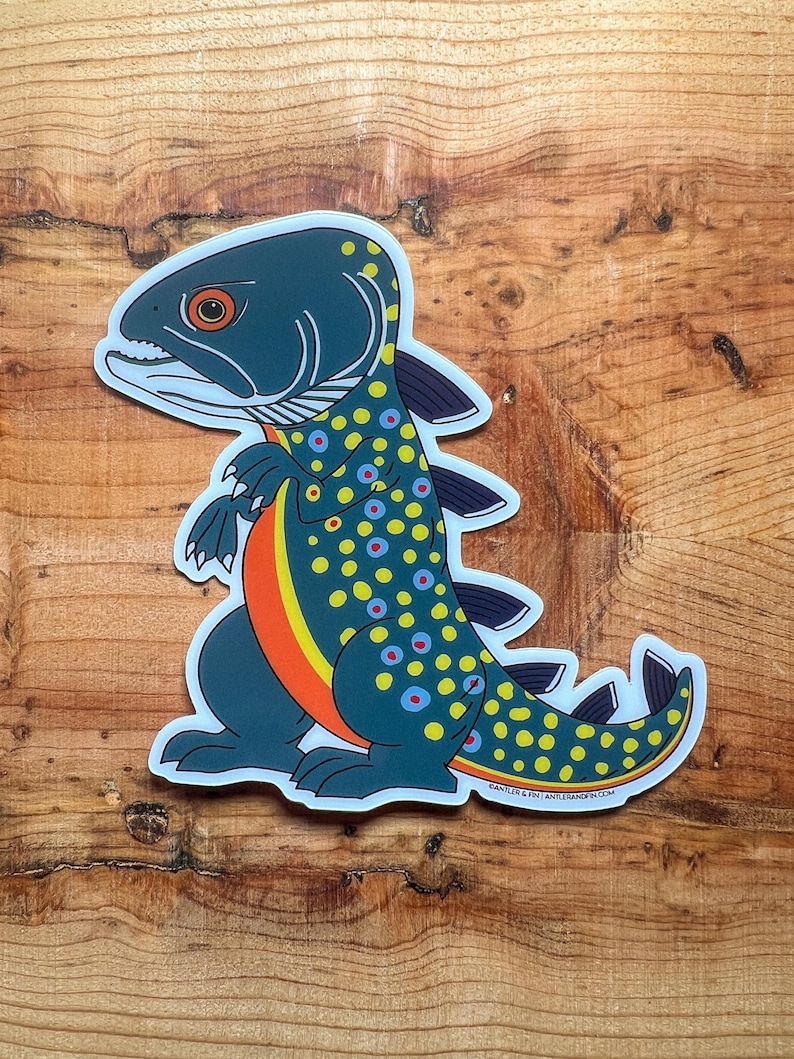 May include: A sticker featuring a cartoon fish-dinosaur hybrid. The creature has a dark teal body with yellow and blue spots, an orange stripe, and a fish-like head. The sticker is on a wooden surface.