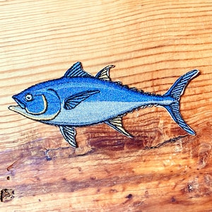 May include: A blue and yellow embroidered patch of a tuna fish on a wooden background.