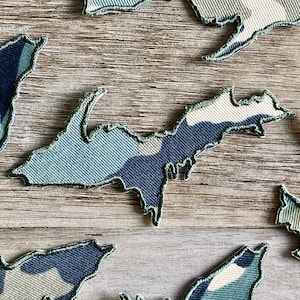 May include: A collection of fabric patches in the shape of the Upper Peninsula of Michigan. The patches feature a camouflage pattern in shades of blue, green, and white, with a green stitched border. The patches are arranged on a wooden surface.