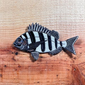 May include: A black and white striped fish with a white body and black stripes. The fish is embroidered on a brown wooden surface.