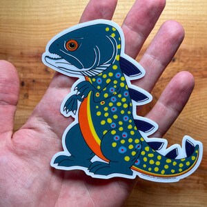 May include: A die-cut sticker featuring a whimsical fish-dinosaur hybrid. The creature has a dark teal body with yellow and blue spots, an orange belly, and a white outline. The sticker is held in a person's hand against a wooden background.