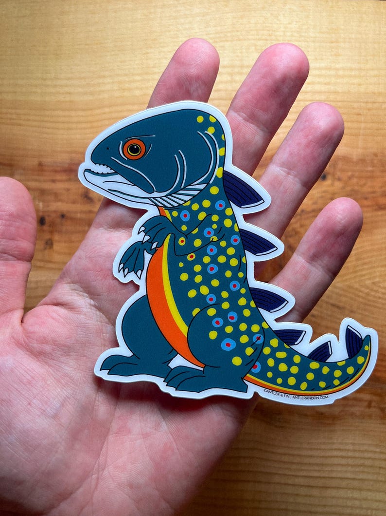 May include: A die-cut sticker featuring a whimsical fish-dinosaur hybrid. The creature has a dark teal body with yellow and blue spots, an orange belly, and a white outline. The sticker is held in a person's hand against a wooden background.