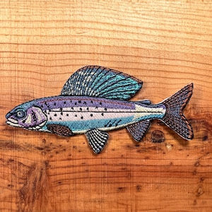 May include: A colorful embroidered patch of a trout fish with purple, blue, and white details. The patch is on a wooden background.