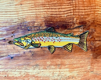 May include: A yellow, green, and white embroidered patch of a trout fish on a wooden surface.