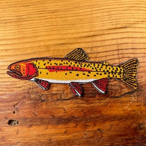 May include: A yellow and orange embroidered patch of a trout fish with black spots and red fins.