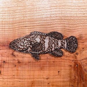 May include: A brown and white embroidered patch of a grouper fish on a wooden background.