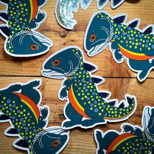 May include: A collection of whimsical stickers featuring a fish-like creature with a dinosaur-like body. The stickers are primarily teal with yellow, orange, and purple accents. The stickers are on a wooden surface.