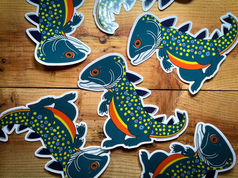May include: A collection of whimsical stickers featuring a fish-like creature with a dinosaur-like body. The stickers are primarily teal with yellow, orange, and purple accents. The stickers are on a wooden surface.