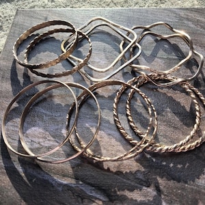 May include: A collection of silver bangles in various designs. The bangles include twisted, hammered, and geometric shapes. The bracelets are displayed on a textured, gray surface, with sunlight casting shadows across the jewelry.
