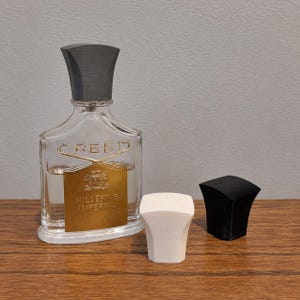 Peut inclure: Un flacon de parfum en verre transparent avec une étiquette dorée portant l'inscription CREED MILLESIME IMPERIAL, surmonté d'un bouchon gris foncé. À côté du flacon se trouvent deux bouchons de remplacement, un blanc et un noir, tous évasés.