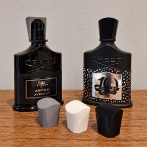 Könnte beinhalten: Zwei schwarze Glasparfümflaschen mit Verschlusskappen, eine mit der Aufschrift Absolu Aventus und die andere 10th Anniversary Aventus. Drei kleine, geometrische, 3D-gedruckte Kappen in Grau, Weiß und Schwarz stehen vor den beiden Parfümflaschen.