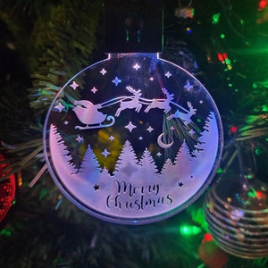 Könnte beinhalten: Ein runder, transparenter Weihnachtsbaumschmuck mit einer weißen Silhouette von Weihnachtsmann in einem Schlitten, der von Rentieren gezogen wird, die über einen verschneiten Wald fliegen. Der Schmuck hat den Text "Merry Christmas" unten.