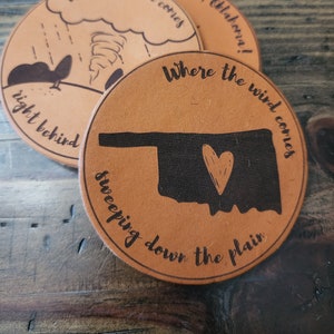May include: Two brown leather coasters with the text "Where the wind comes sweeping down the plain" and a silhouette of the state of Oklahoma with a heart in the center. The other coaster has the text "When the wind comes right behind" and a cartoon of a tornado.