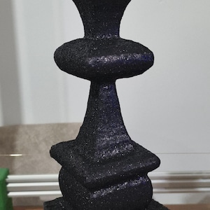 May include: A black, textured chess piece, possibly a queen, is displayed. It features a square base, a cylindrical middle, and a crown-shaped top. The piece is set on a wooden surface.