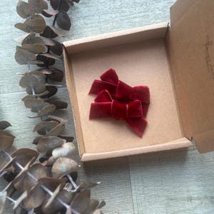 May include: Three red velvet hair bows in a small cardboard box. The bows are arranged in a stack, with the largest bow on top.