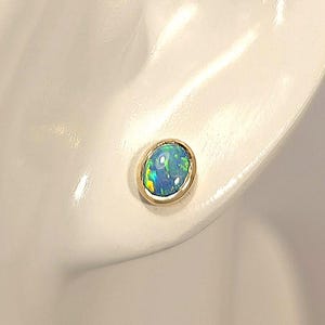 May include: A single gold stud earring with an oval, iridescent opal stone. The opal displays shades of blue, green, and yellow. The earring is set in a simple gold bezel, showcasing the stone's natural beauty. The earring is displayed on a white mannequin.