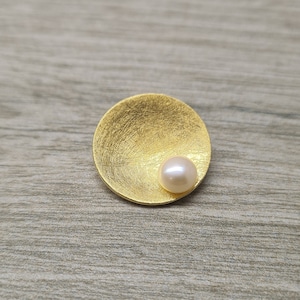 May include: A gold-toned disc with a textured surface and a single white pearl in the center.