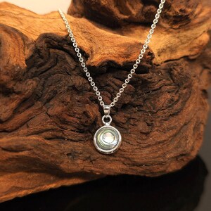 May include: A silver necklace with a circular pendant. The pendant features a pearlescent, iridescent design within a silver frame. The chain is delicate and hangs against a textured, dark brown wooden background.