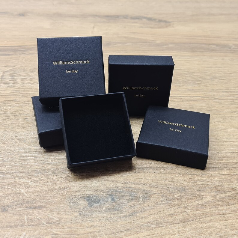 May include: Several small, black jewelry boxes are arranged on a wooden surface. The boxes have a square shape and a matte finish. The text "WilliamsSchmuck bei Etsy" is printed in gold on the top of each box.