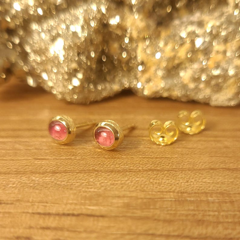Puede incluir: Un par de pendientes de oro con piedras preciosas rosas. Los pendientes est&aacute;n sobre una superficie de madera.
